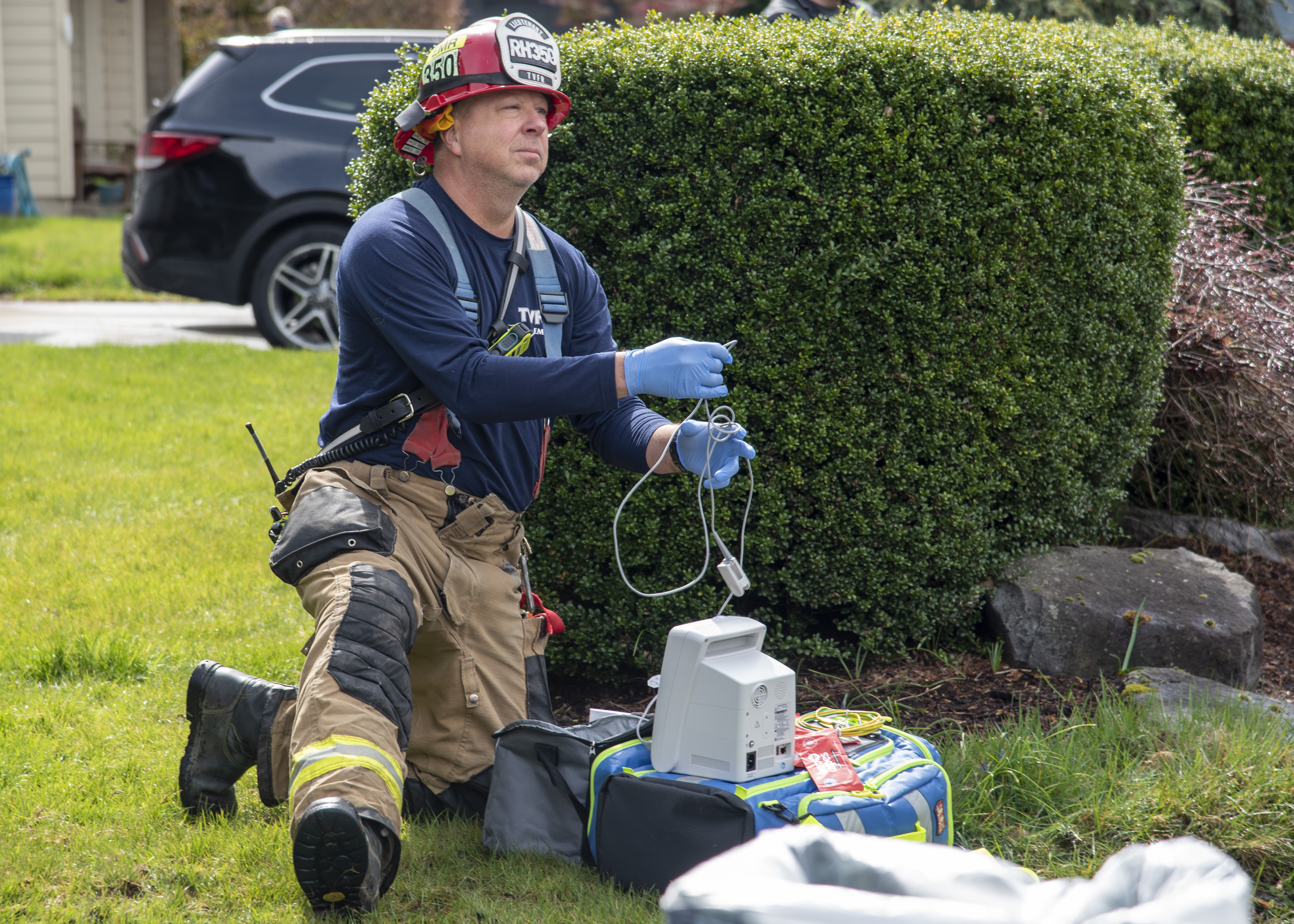 Volunteer kneels on ground holding medical device to check firefighter's vitals at fire scene
